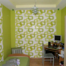 Pok&oacute;j dziecięcy z zielonymi ścianami i tapetą w geometryczne wzory, drewniana podłoga, biurko z komputerem i szafa z jasnego drewna.