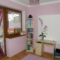 Pokój w odcieniach różu z drewnianymi oknami i podłogą, wyposażony w biurko, regał z książkami zwieńczony kapeluszem, dekoracyjną lampę, zasłony i dywan w podobnej tonacji kolorystycznej.