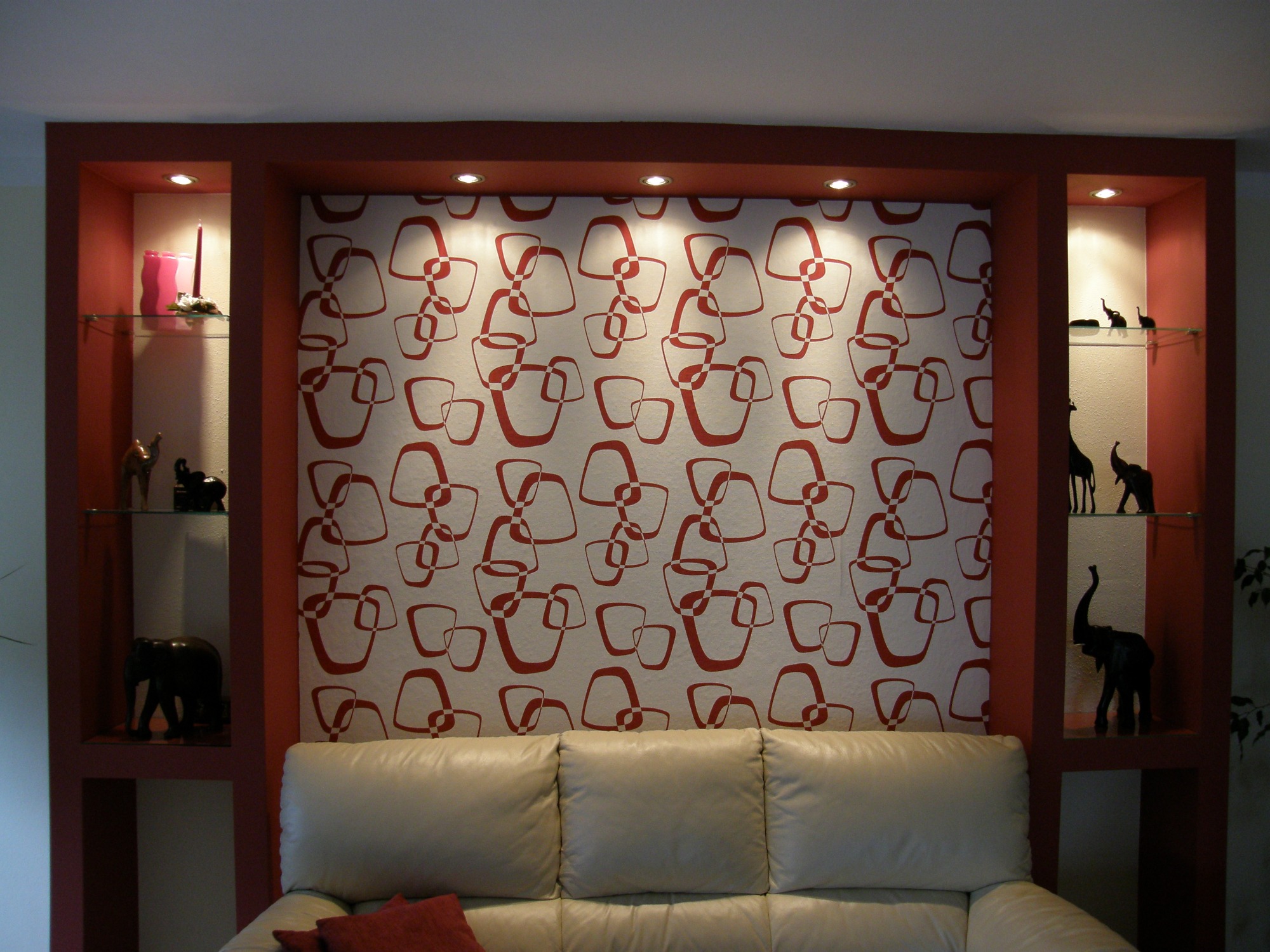 Ściana z wnękami pomalowanymi na czerwono, z podświetlanym wzorzystym tłem i dekoracyjnymi figurkami słoni na szklanych półkach, poniżej kremowa sofa.
