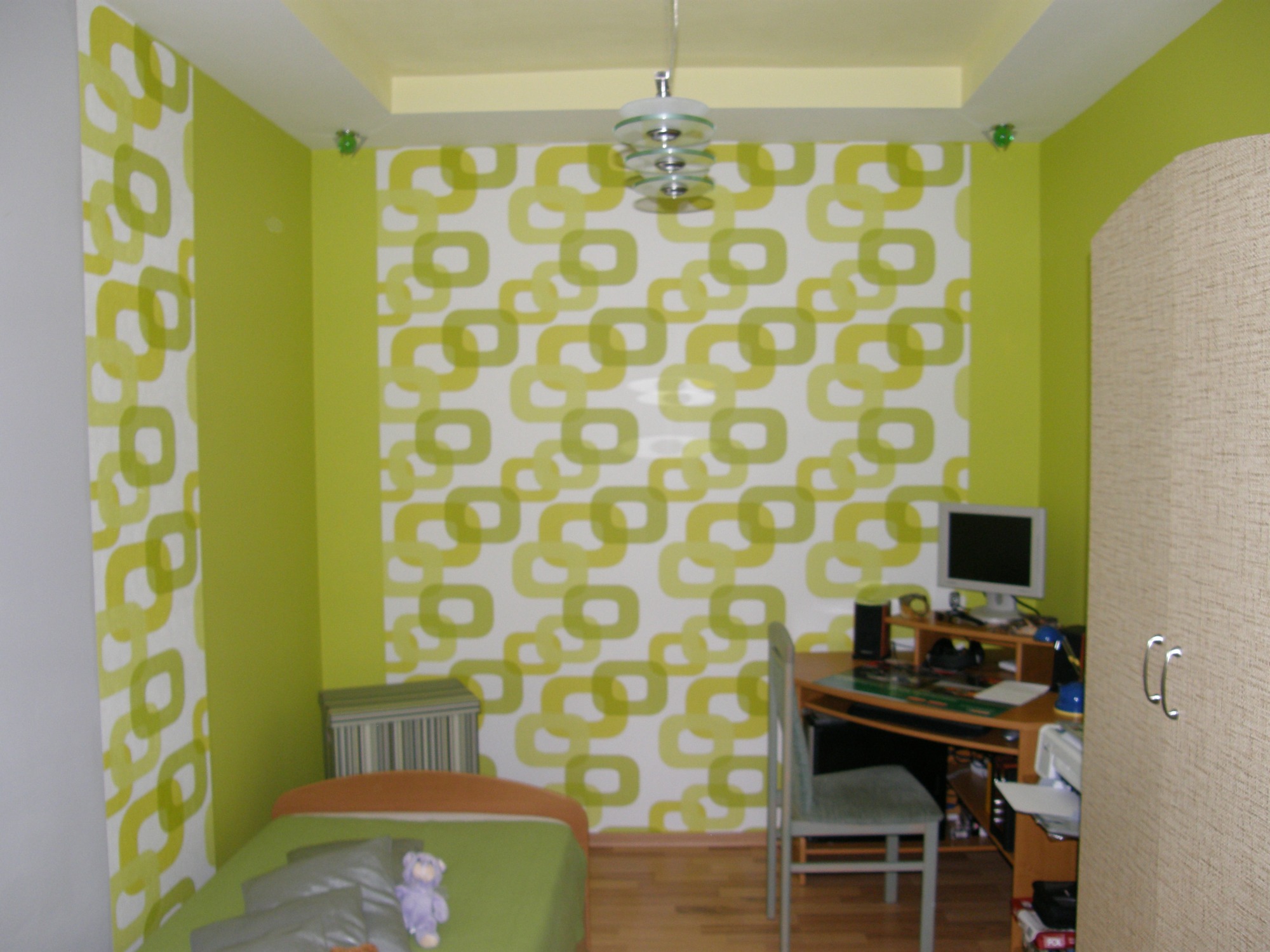 Pokój dziecięcy z zielonymi ścianami i tapetą w geometryczne wzory, drewniana podłoga, biurko z komputerem i szafa z jasnego drewna.