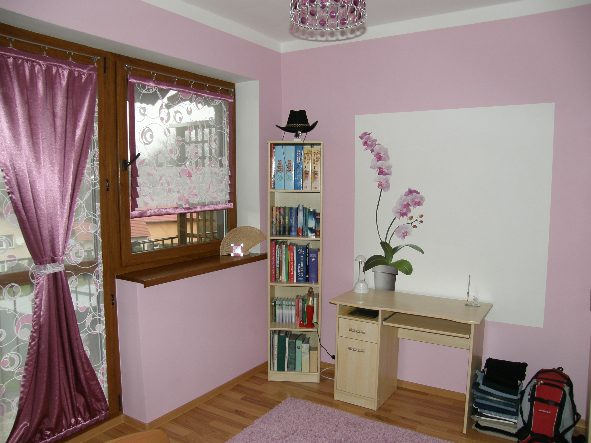 Pokój w odcieniach różu z drewnianymi oknami i podłogą, wyposażony w biurko, regał z książkami zwieńczony kapeluszem, dekoracyjną lampę, zasłony i dywan w podobnej tonacji kolorystycznej.