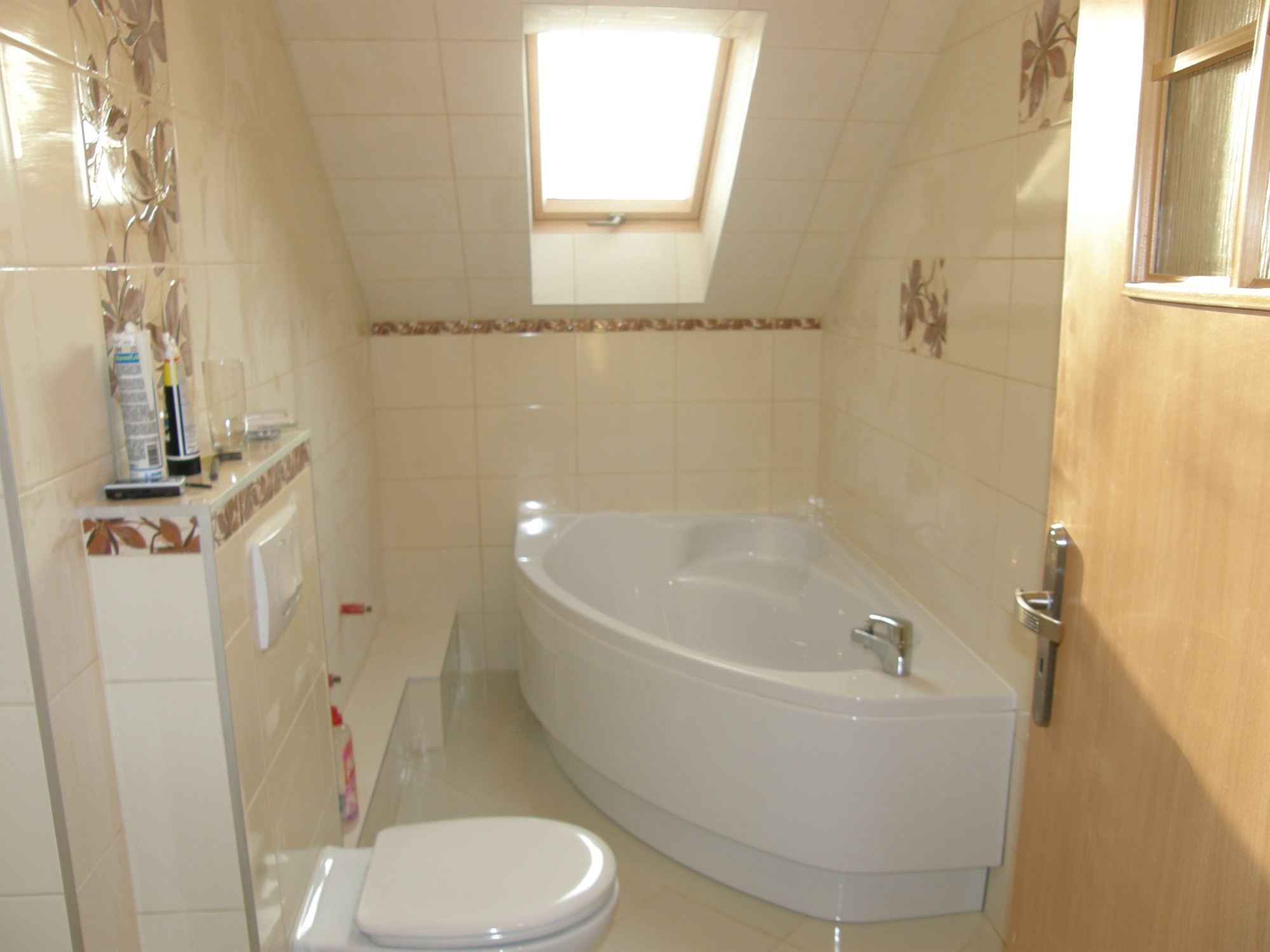 Jasna łazienka na poddaszu w Ostrożnicy z narożną wanną, beżowymi płytkami z motywem kwiatowym i oknem dachowym.