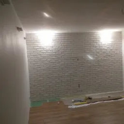 Wnętrze pokoju w trakcie prac wykończeniowych: ściana wyłożona dekoracyjną białą cegłą, częściowo ułożona podłoga z paneli, narzędzia i materiały budowlane na folii ochronnej.