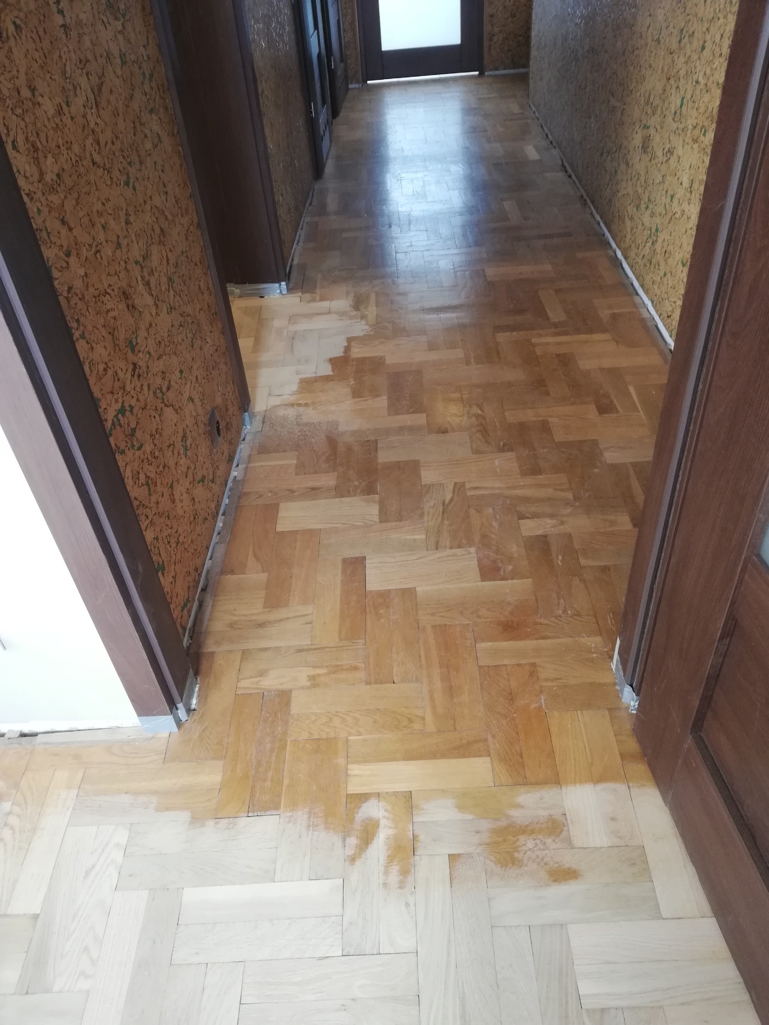 Parkiet ułożony w jodełkę, częściowo wyszlifowany, z widocznym kontrastem między starą a odnowioną powierzchnią w korytarzu z korkowymi ścianami.