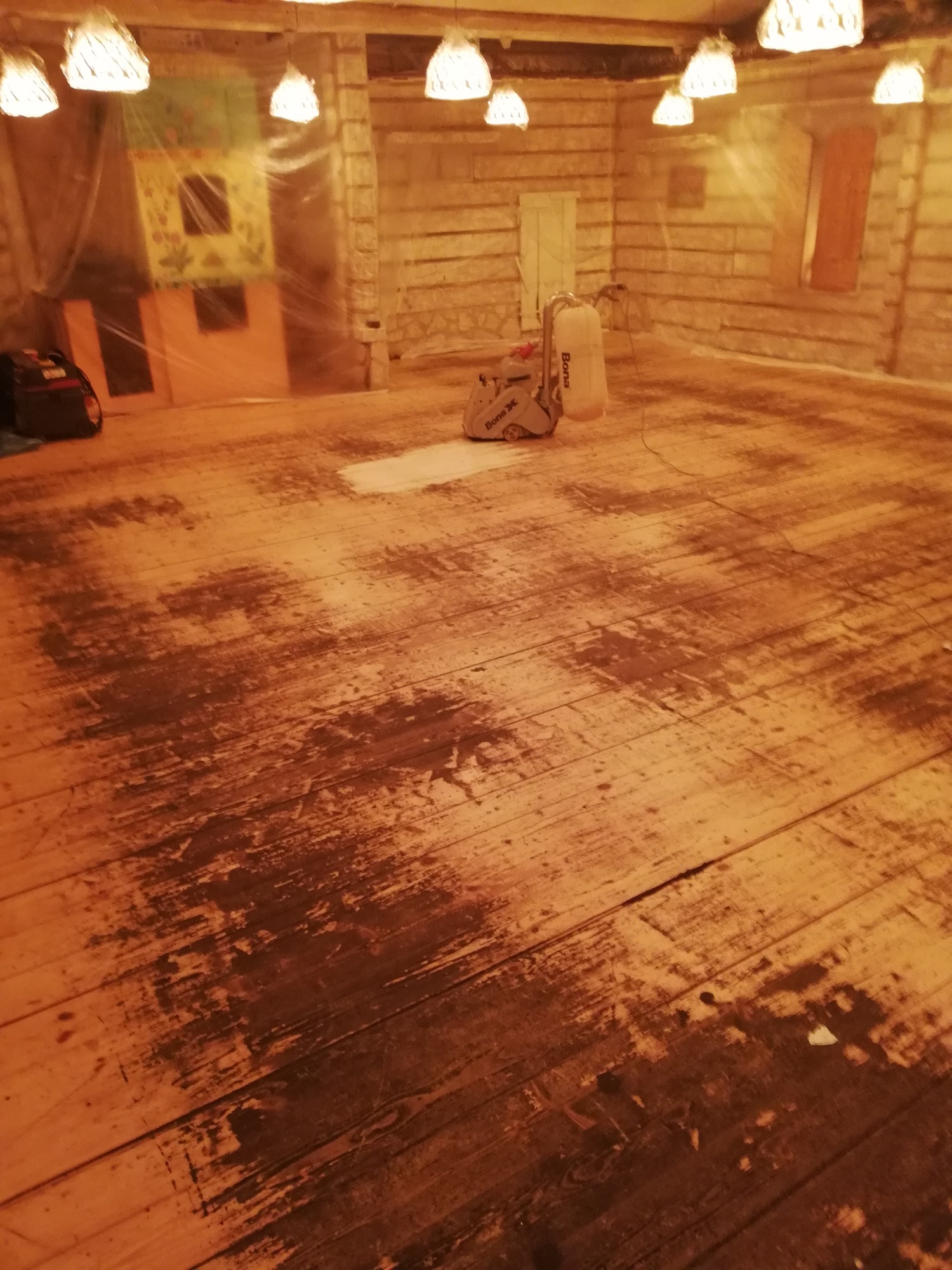 Częściowo wyszlifowana drewniana podłoga w pomieszczeniu z drewnianymi ścianami, widoczna maszyna do szlifowania Bona X i zasłonięty folią dziecięcy domek.