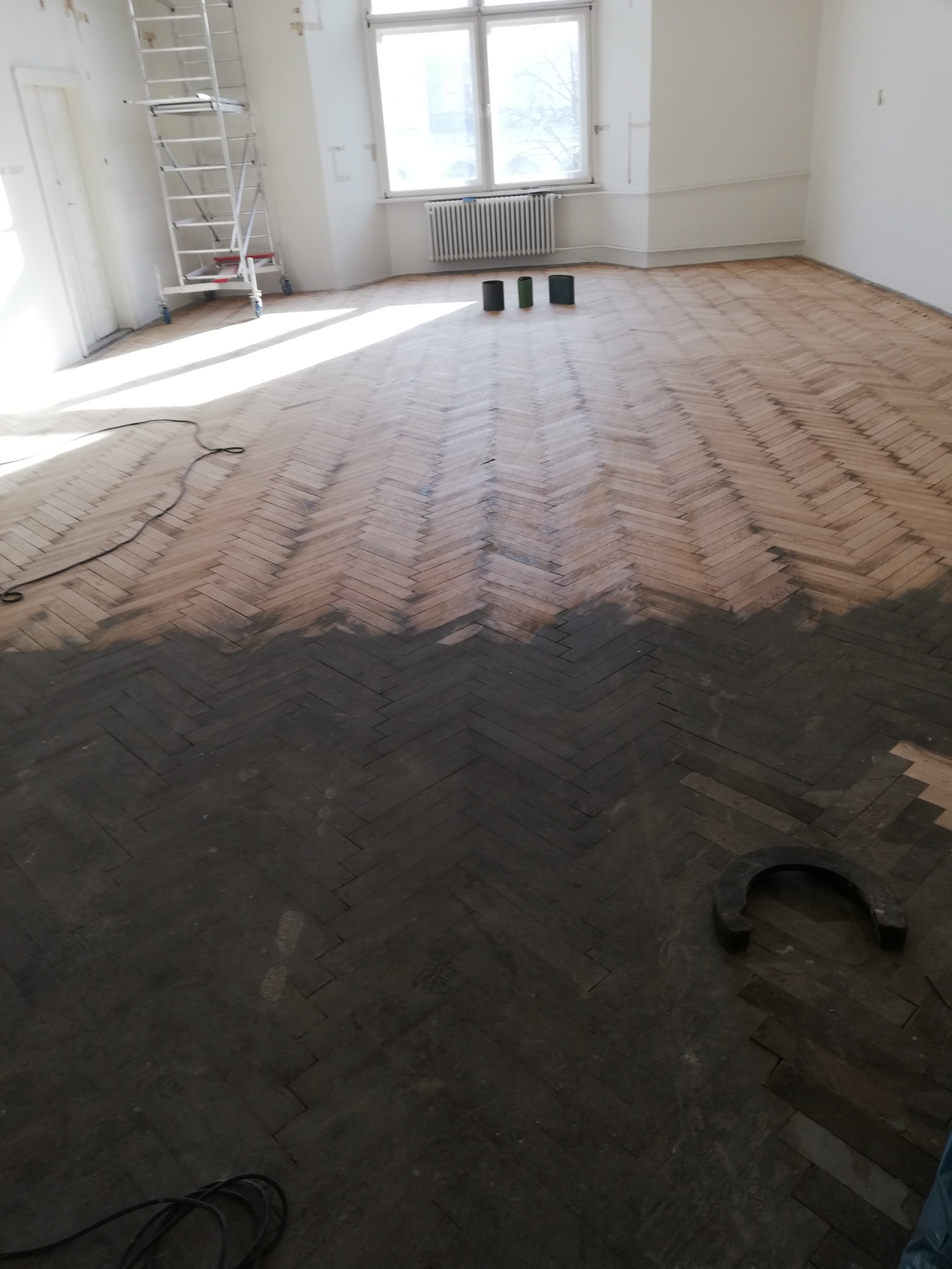 Parkiet w jodełkę częściowo pokryty ciemną bejcą, widoczne trzy puszki z bejcą przy oknie i drabina malarska w tle