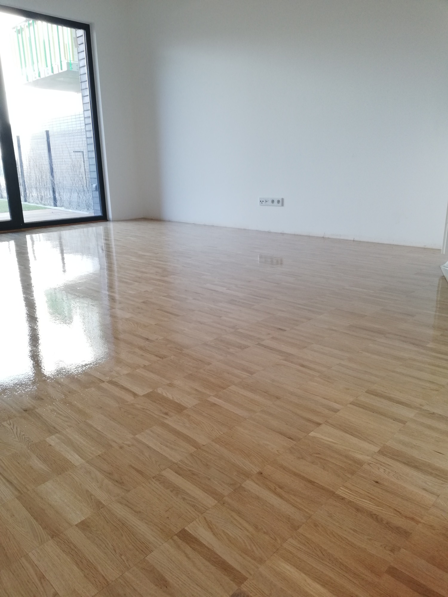 Błyszcząca, świeżo polakierowana parkietowa podłoga w pustym pokoju z dużym oknem balkonowym i białymi ścianami.