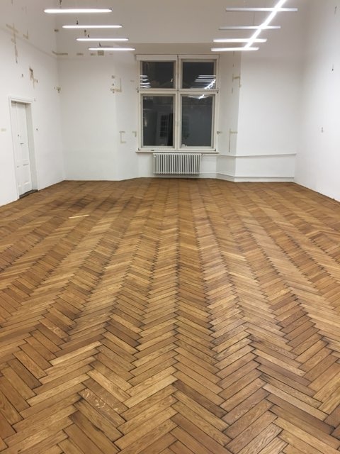 Parkiet ułożony w jodełkę po renowacji w pustym pokoju z oknem, widoczne ślady po taśmach malarskich na ścianach.