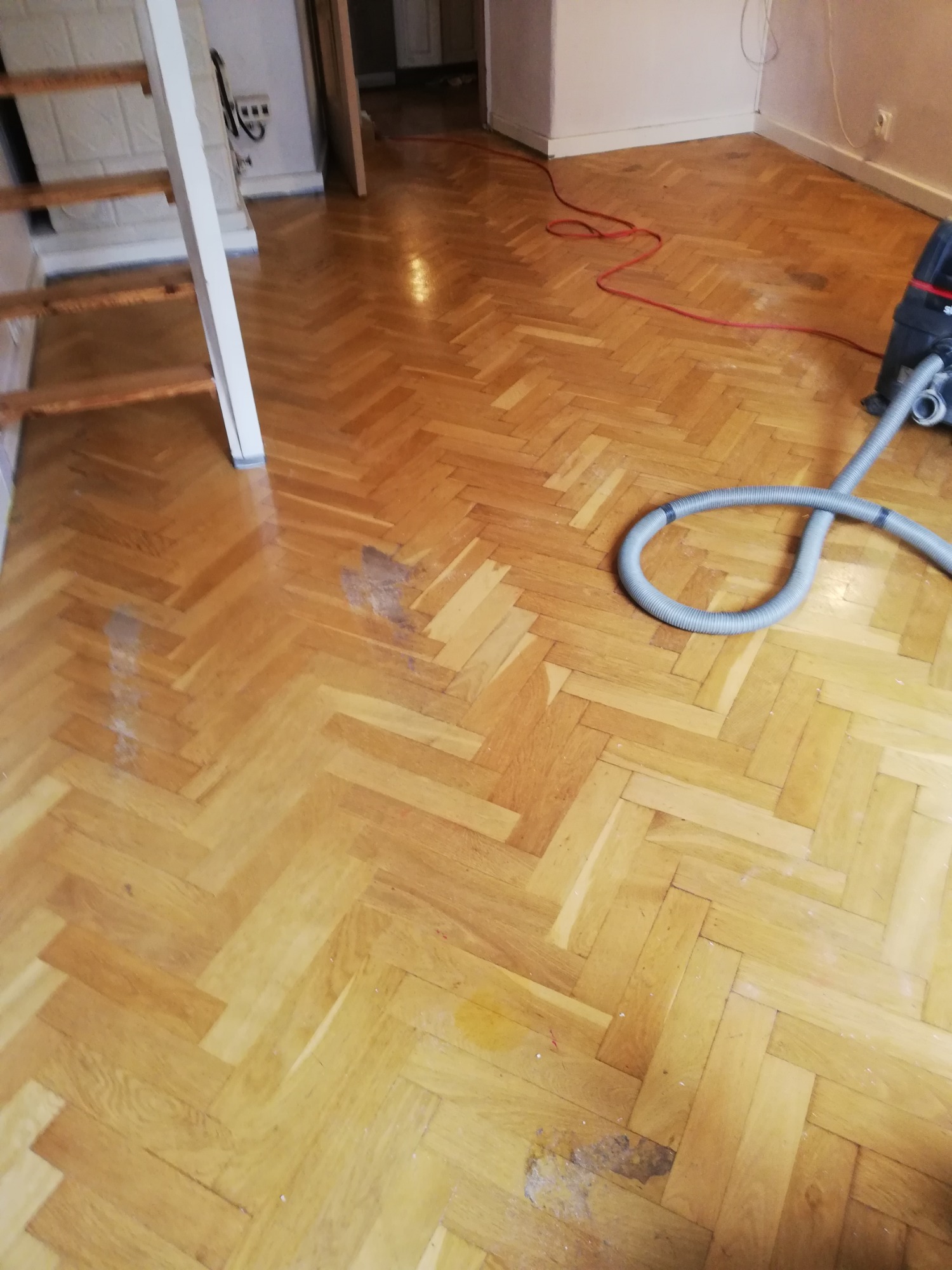 Parkiet ułożony w jodełkę przed cyklinowaniem, widoczne plamy i zabrudzenia, odkurzacz przemysłowy w rogu pomieszczenia, schody w tle.