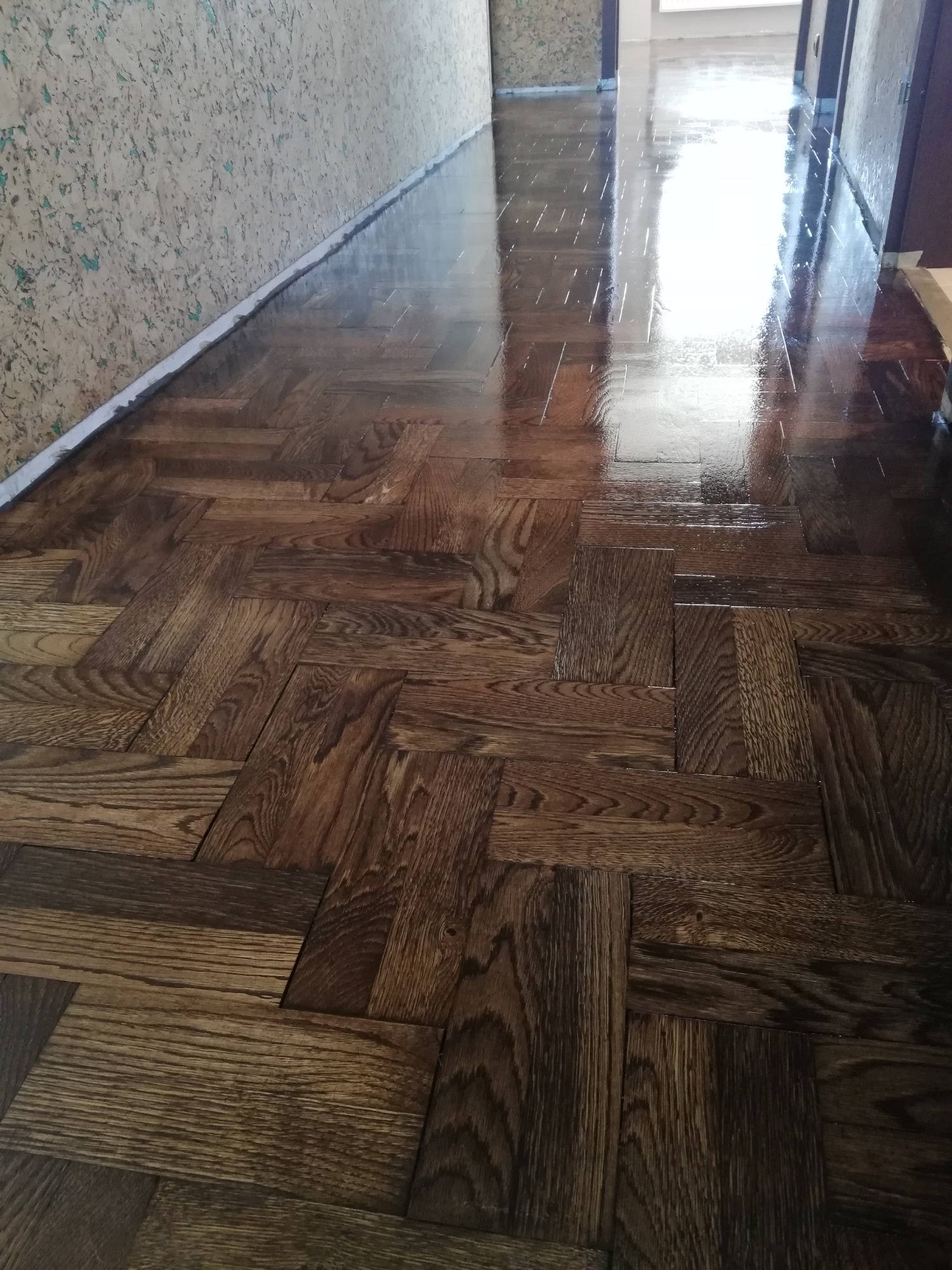 Błyszcząca, świeżo polakierowana podłoga parkietowa ułożona we wzór jodełki francuskiej, widoczna po zabiegu renowacji, z jasnym odbiciem światła w tle na końcu korytarza.