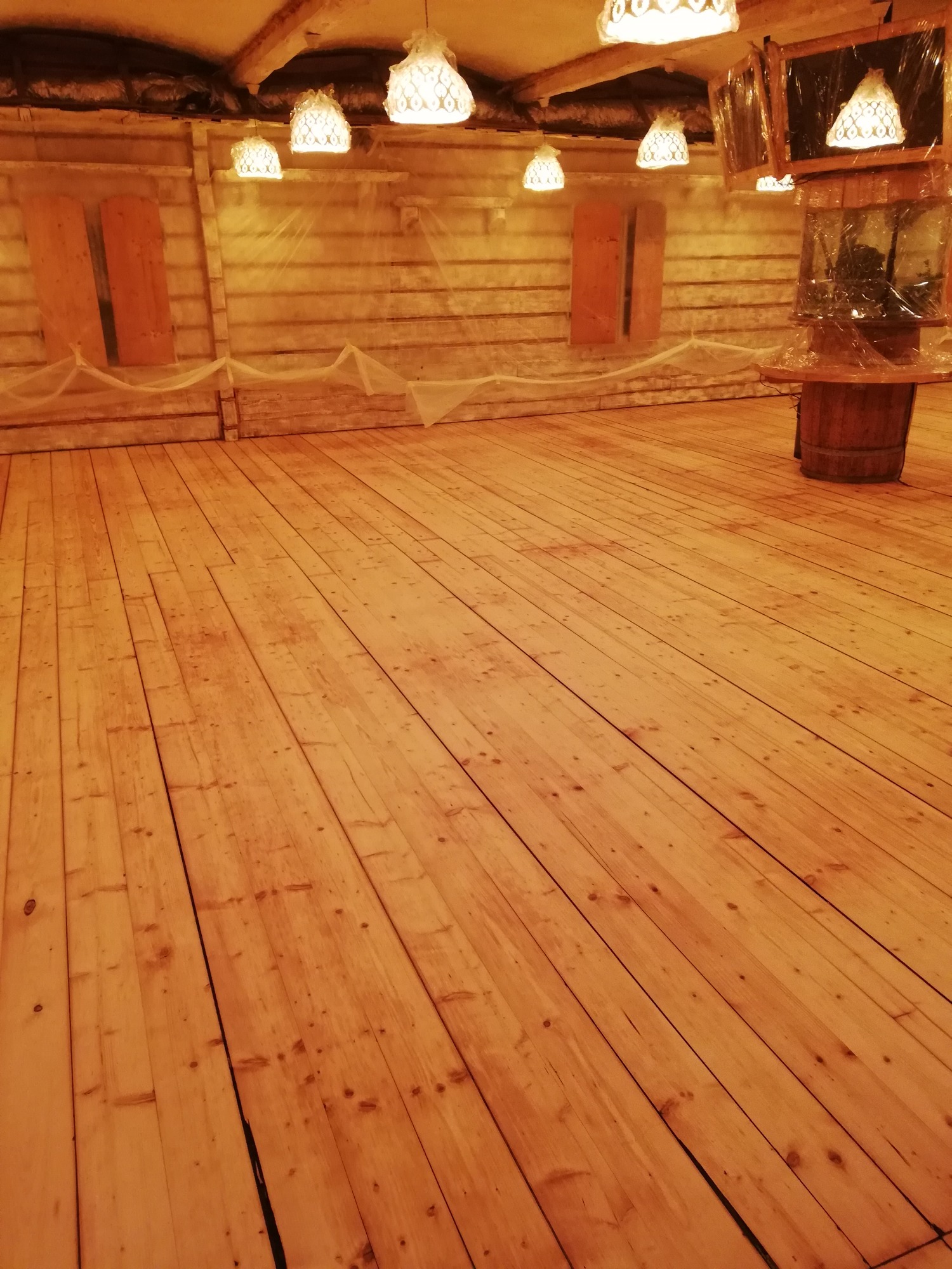 Wycyklinowana, jasna podłoga z desek w pomieszczeniu z drewnianymi ścianami i dekoracyjnymi lampami. Podłoga jest gładka i ma widoczne słoje drewna.