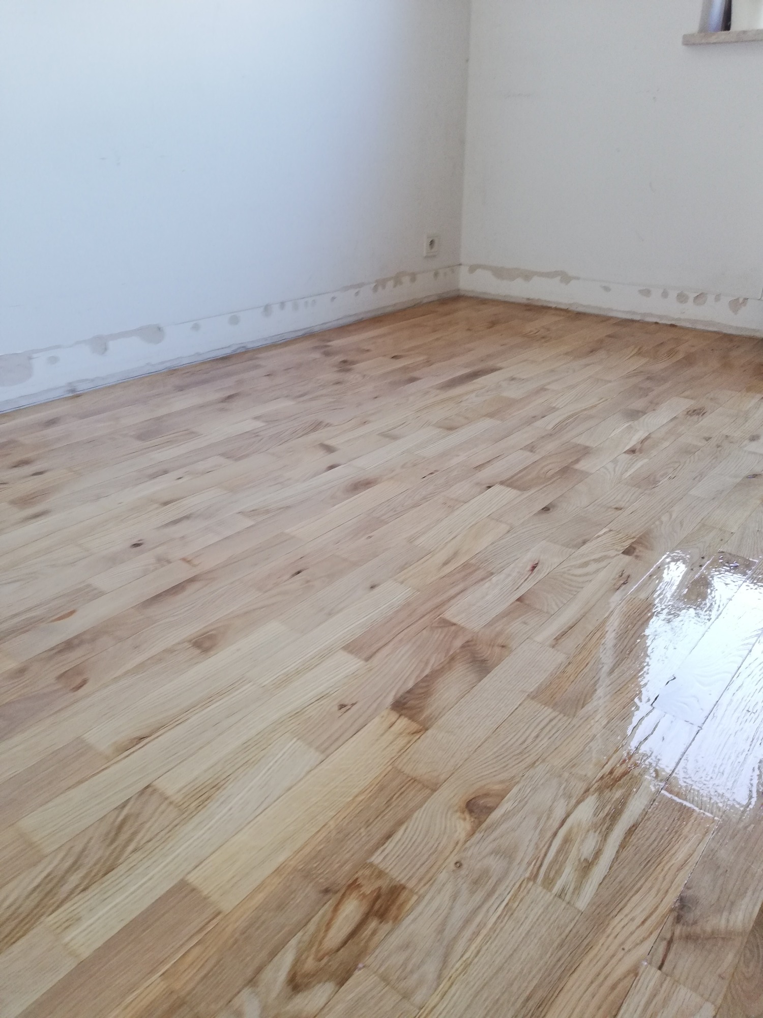 Parkiet po nałożeniu lakieru, widoczne odbicie światła na mokrej powierzchni, ściany przygotowane do malowania z widocznymi śladami po taśmie malarskiej przy podłodze.