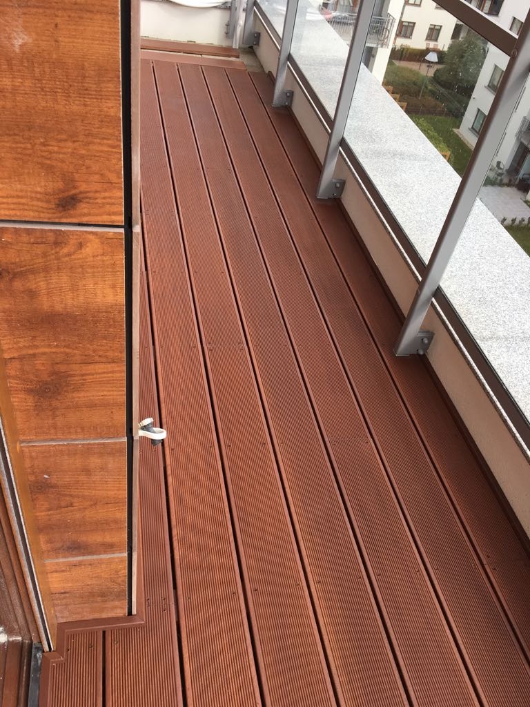 Balkon z desek kompozytowych w odcieniach brązu, widok z góry, balustrada szklana z metalowymi wspornikami, widok na otoczenie budynku.