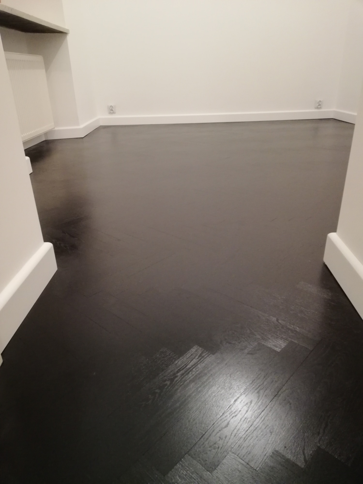 Wypolerowana, ciemna podłoga parkietowa ułożona w jodełkę w pustym, białym pomieszczeniu z listwami przypodłogowymi.