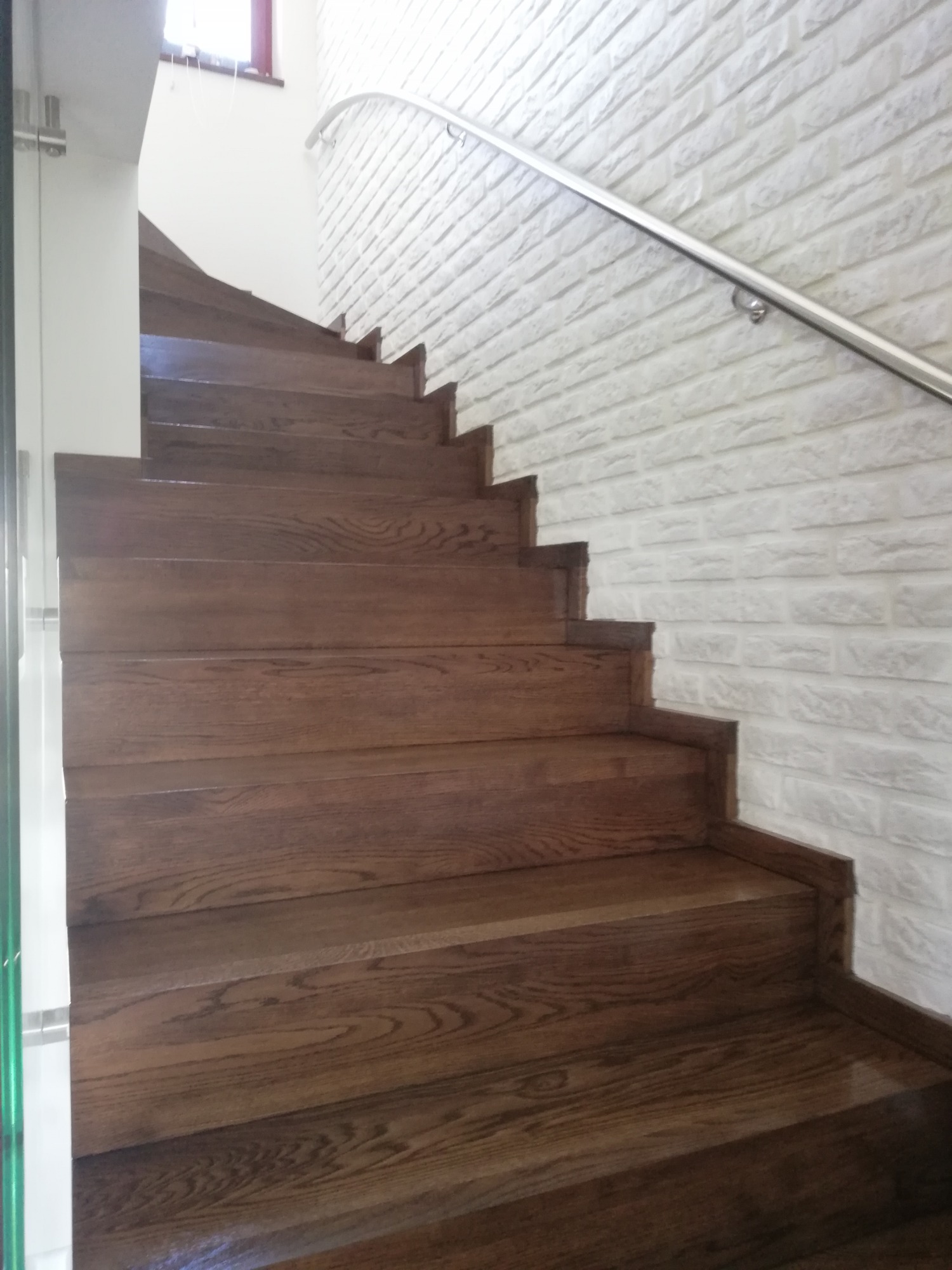 Drewniane schody dębowe z metalową poręczą na tle ściany z białej cegły dekoracyjnej, widoczne detale usłojenia drewna i mocowania poręczy.