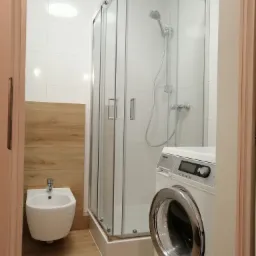 Nowoczesna łazienka w Gliwicach z kabiną prysznicową, bidetem, pralką Miele i drewnopodobnymi płytkami na podłodze i części ściany, widok zza futryny drzwi.