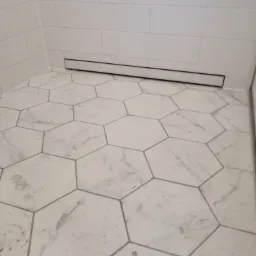 Wykończona podłoga w łazience z białych heksagonalnych płytek imitujących marmur, z widocznym odpływem liniowym przy ścianie i fragmentem kabiny prysznicowej.