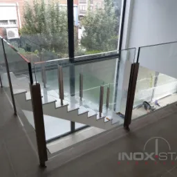 Nowoczesne schody z białymi stopniami i szklaną balustradą z metalowymi słupkami w jasnym wnętrzu, widok z góry z logo firmy w prawym dolnym rogu.