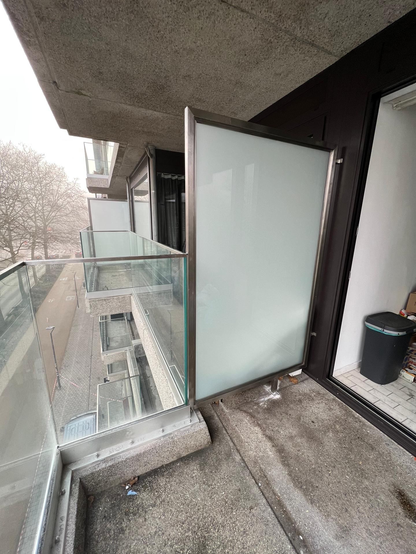 Balkon z szklaną balustradą i przesuwną osłoną, widok z góry na ulicę w pochmurny dzień. W tle budynki mieszkalne i drzewa bez liści. Na balkonie widoczny kosz na śmieci.