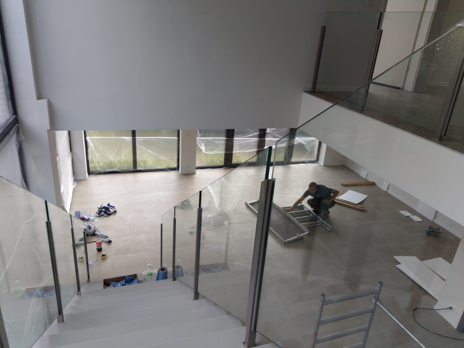 Montaż szklanej balustrady w nowoczesnym domu. Widok z góry na schody z białymi stopniami i szklanymi panelami. Pracownik na kolanach przykłada panel do mocowania. Okna zabezpieczone folią ochronną.