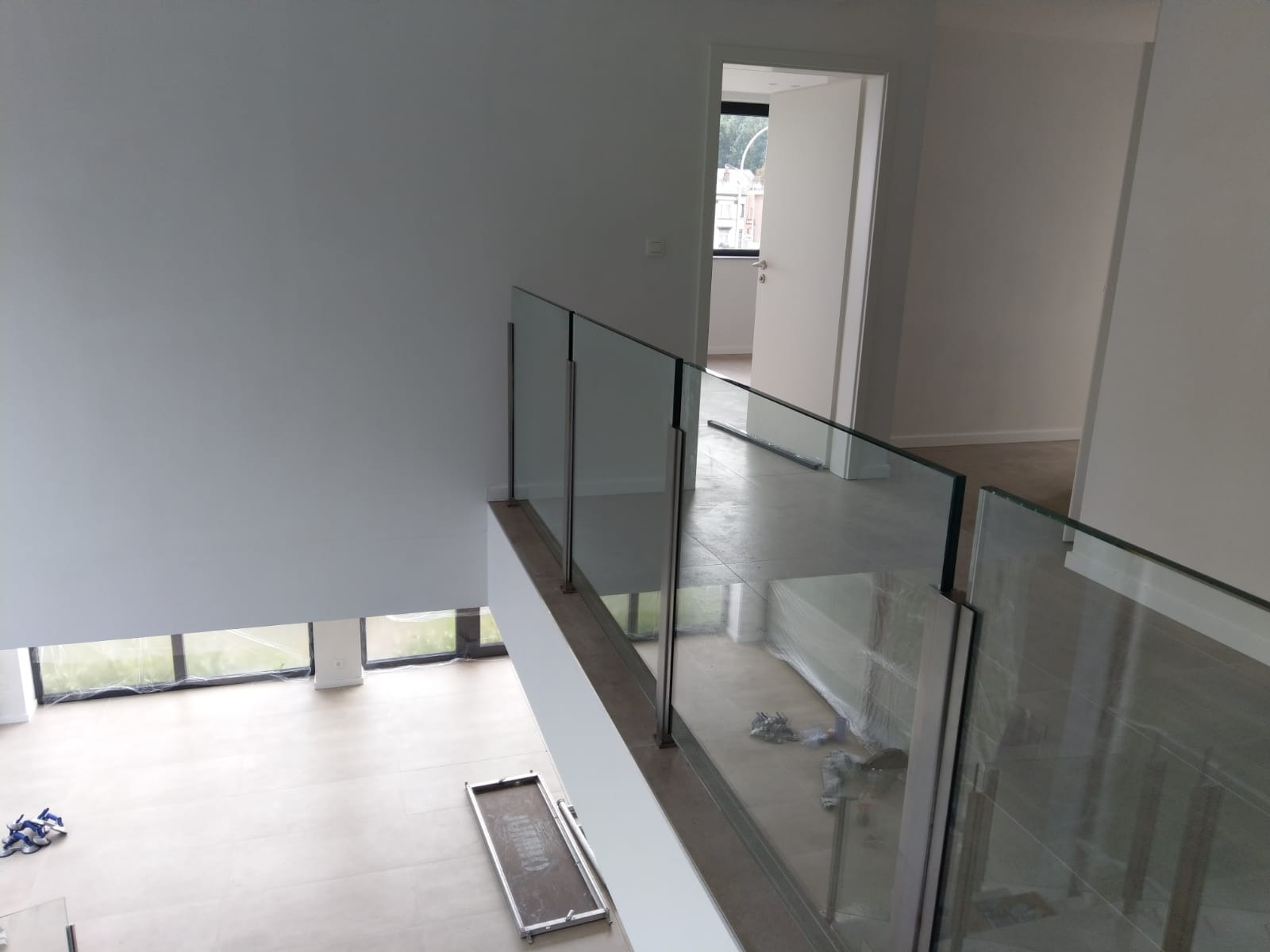 Wnętrze domu w trakcie wykańczania z zamontowaną szklaną balustradą z metalowymi słupkami, widok z góry na parter z dużymi oknami.