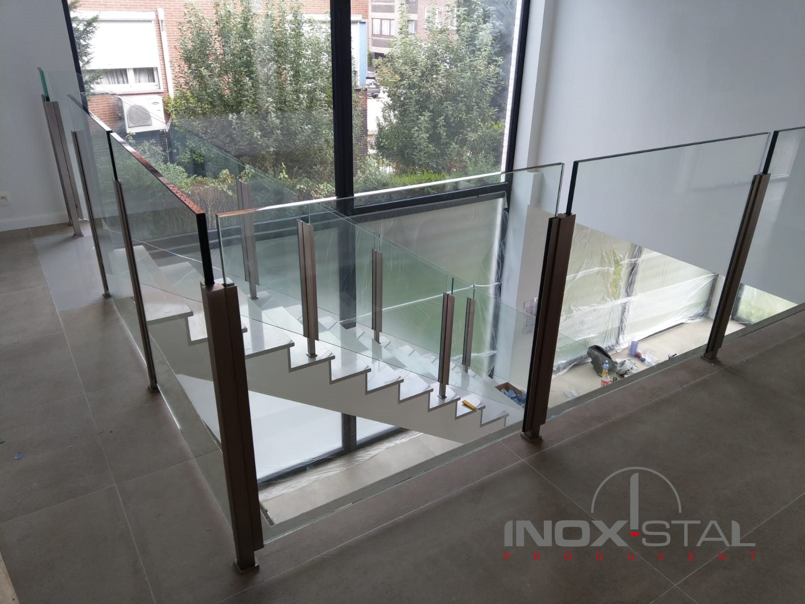 Nowoczesne schody z białymi stopniami i szklaną balustradą z metalowymi słupkami w jasnym wnętrzu, widok z góry z logo firmy w prawym dolnym rogu.