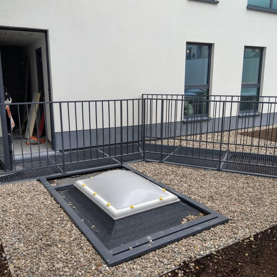 Zewnętrzna balustrada z metalu w kolorze antracytowym, otaczająca okno dachowe na poziomie gruntu, wykończone drobnym żwirem, na tle białej ściany budynku z oknami.