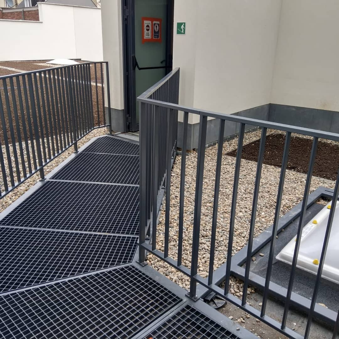 Metalowa konstrukcja z kratownicą i szarymi barierkami prowadząca do drzwi z piktogramami na zewnątrz budynku.