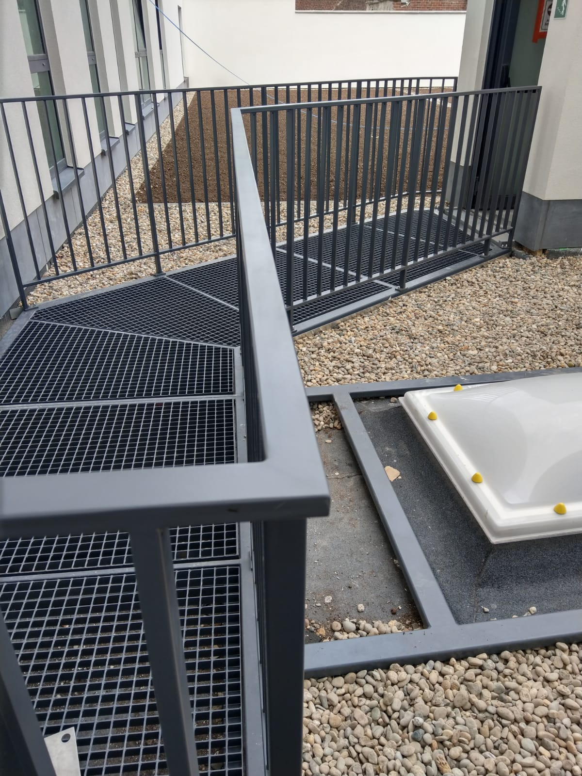 Metalowa konstrukcja schodów kratowych z szarą poręczą na zewnątrz budynku, otoczona drobnymi kamieniami i z widocznym świetlikiem dachowym z żółtymi elementami.