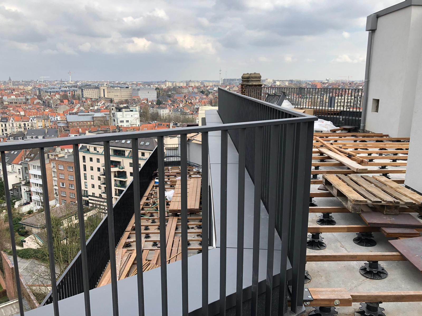 Montaż balustrady na tarasie na dachu budynku z widokiem na miasto, widoczne elementy konstrukcji tarasu z regulowanymi wspornikami i drewnianymi legarami.