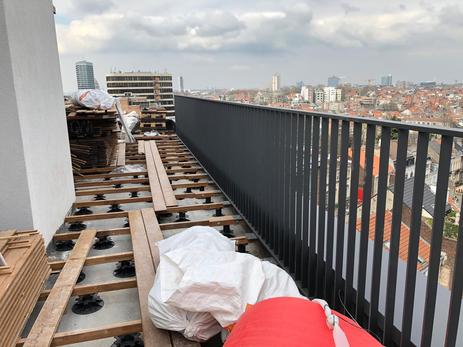 Montaż drewnianego tarasu na dachu budynku z widokiem na miasto, szara barierka ochronna w tle, materiały budowlane w workach i deski na podkładkach regulowanych.