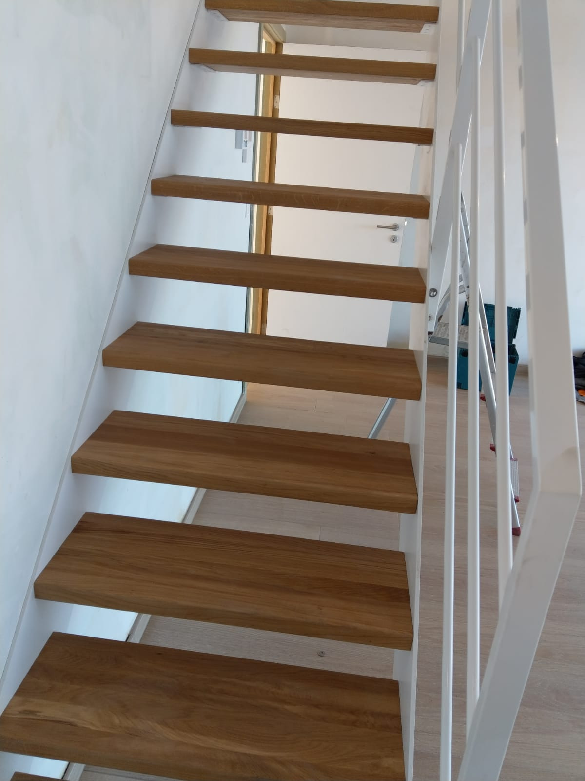 Nowoczesne schody z drewnianymi stopniami i białą metalową konstrukcją oraz poręczą, widok z dołu, w tle jasne ściany i drzwi.