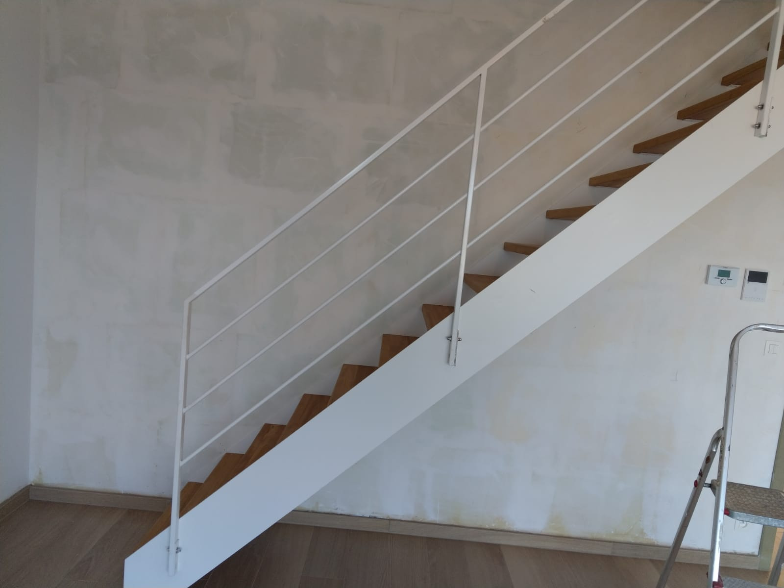 Białe schody metalowe z drewnianymi stopniami i minimalistyczną, białą balustradą z poziomych prętów na tle ściany w trakcie malowania, widoczna drabina malarska.