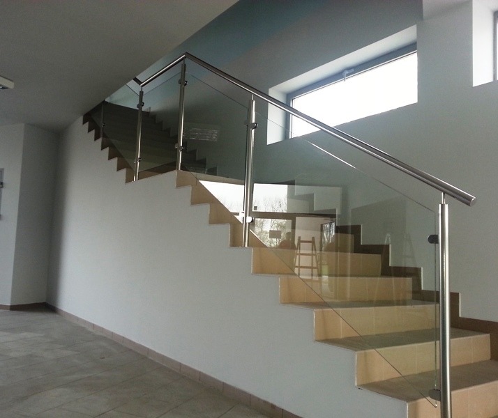 Schody z beżowymi stopniami zabezpieczone szklaną balustradą z metalową poręczą, widok z dołu na jasne wnętrze.