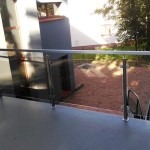 Szklana balustrada balkonowa z nierdzewnymi słupkami, widok z bliska na elementy mocujące, tło stanowi zielony ogród.