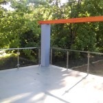 Zewnętrzna szklana barierka z metalowymi słupkami i poręczą, zamontowana na betonowym tarasie z widokiem na drzewa.