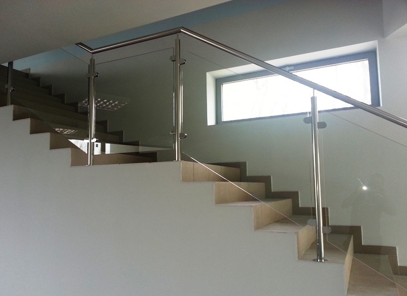 Szklana balustrada ze słupkami ze stali nierdzewnej zamontowana na schodach z jasnych płytek, widok z boku.