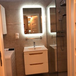 UO Mariusz Baron - Nowoczesna łazienka z podświetlanym lustrem, kabiną prysznicową typu walk-in i umywalką z szafką w kolorze białym, widok zza drzwi.