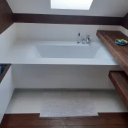 Minimalistyczna łazienka z białą wanną w zabudowie, drewnianymi elementami wykończeniowymi i dywanikiem z mikrofibry