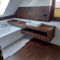 UO Mariusz Baron - Nowoczesna łazienka na poddaszu z drewnianą podłogą i blatem pod umywalkę, biała wanna obok,  dekoracyjna miseczka na blacie, brązowe panele ścienne, okno dachowe.