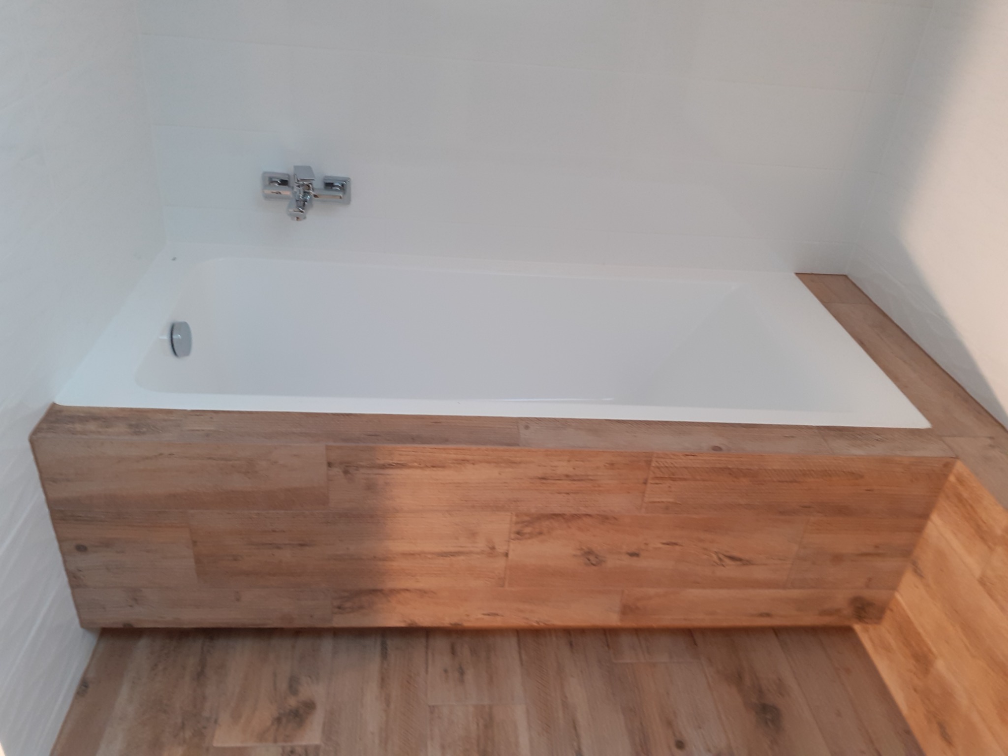 Biała wanna obudowana panelami imitującymi drewno, z chromowaną baterią na białej ścianie pokrytej płytkami.