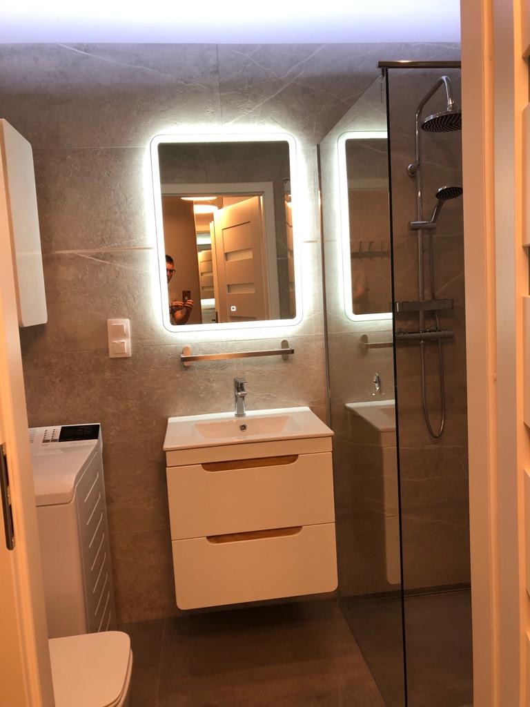 Nowoczesna łazienka z podświetlanym lustrem, kabiną prysznicową typu walk-in i umywalką z szafką w kolorze białym, widok zza drzwi.