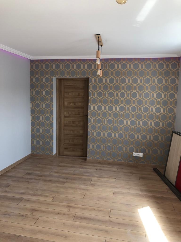 Wykończone wnętrze pokoju z drewnianą podłogą, tapetą w geometryczne wzory i drewnianymi drzwiami; widoczna nowoczesna lampa wisząca z trzema żarówkami.