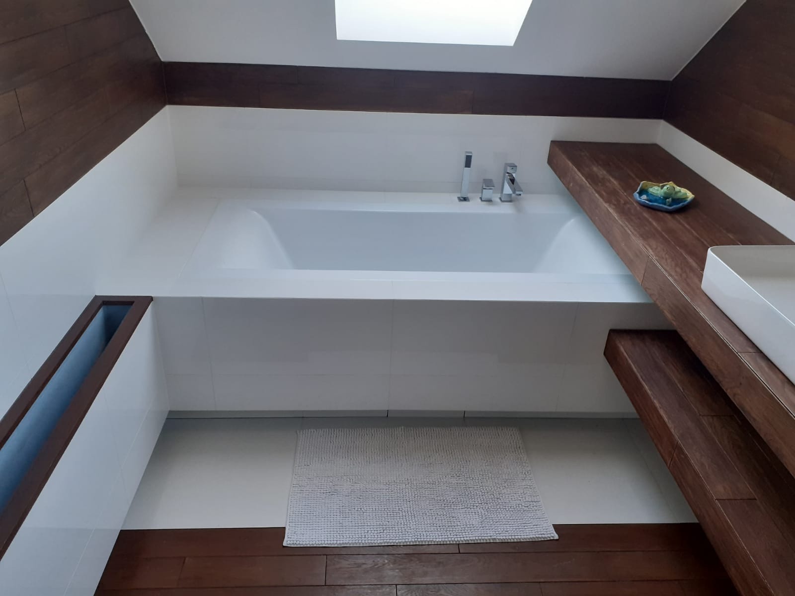 Minimalistyczna łazienka z białą wanną w zabudowie, drewnianymi elementami wykończeniowymi i dywanikiem z mikrofibry