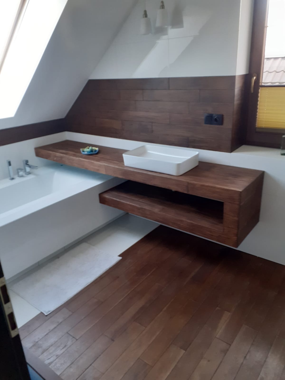 Nowoczesna łazienka na poddaszu z drewnianą podłogą i blatem pod umywalkę, biała wanna obok,  dekoracyjna miseczka na blacie, brązowe panele ścienne, okno dachowe.