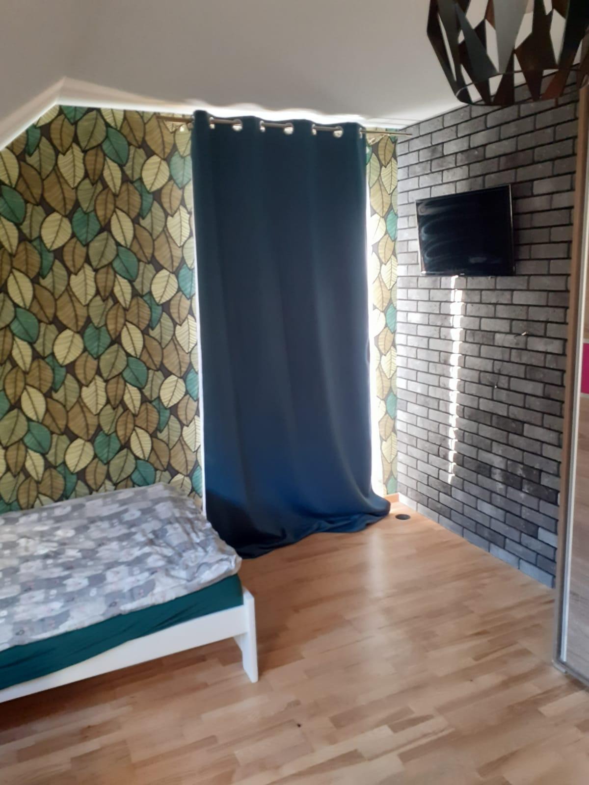 Pokój po odświeżeniu: ściana z szarej cegły z wiszącym telewizorem, zasłona w odcieniach granatu, tapeta w liście oraz drewniana podłoga.
