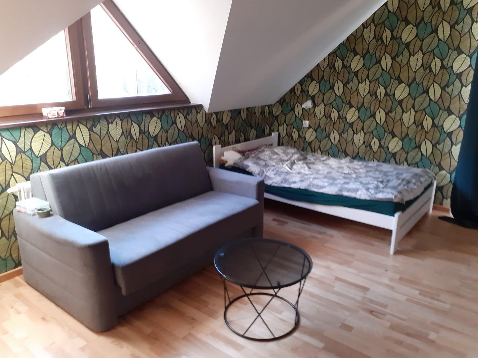 Pokój na poddaszu z tapetą w liście, szarą sofą, okrągłym stolikiem kawowym i łóżkiem z białą ramą.
