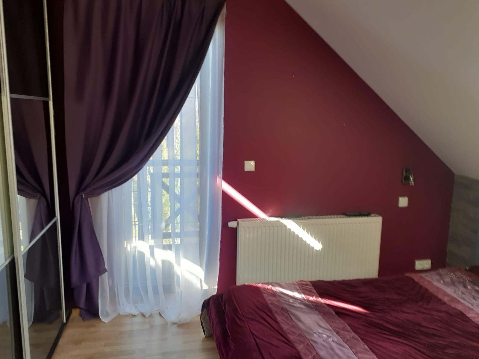 Pokój na poddaszu z bordową ścianą, białym grzejnikiem i częściowo zasłoniętym oknem z białymi firankami oraz ciemnofioletowymi zasłonami, z widocznym fragmentem łóżka z bordową pościelą.