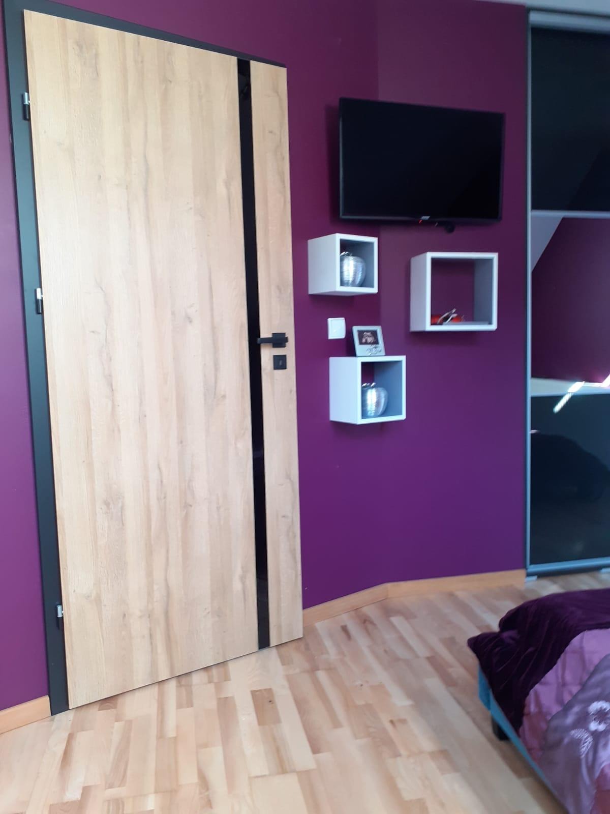 Drzwi wewnętrzne o jasnym, drewnopodobnym wykończeniu z czarnymi akcentami na tle fioletowej ściany z białymi półkami kostkami i telewizorem.