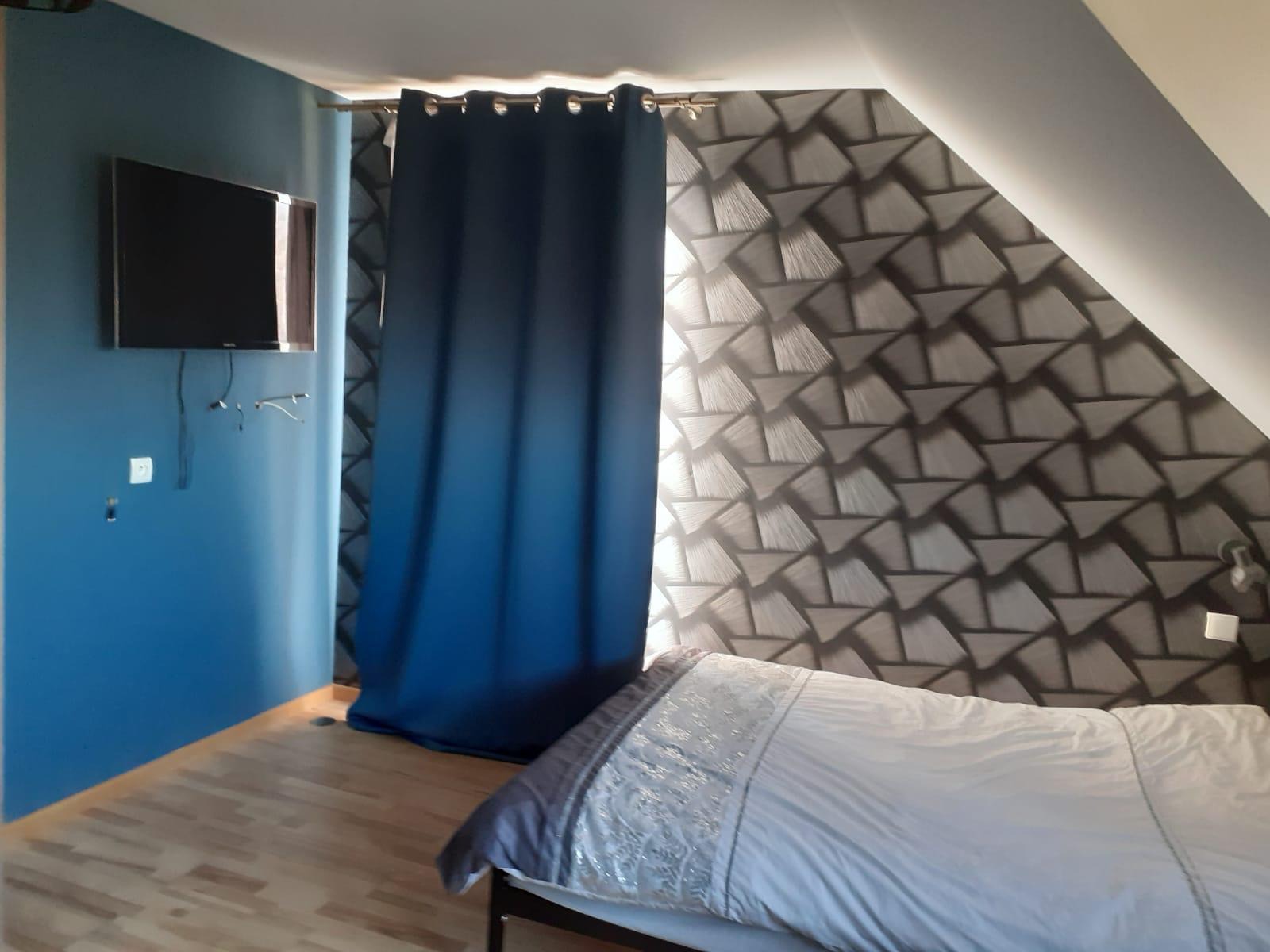 Pokój na poddaszu z niebieską ścianą, telewizorem, wzorzystą tapetą, zasłoną ombre i częściowo widocznym łóżkiem.